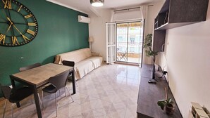 Una habitación pequeña y moderna con una pared de acento verde, un reloj de pared grande, un sofá, una mesa de comedor con sillas y una pequeña cocina con una planta.