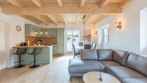A modern kitchen with wooden beams, a dining area with a grey sofa and a small round table, and a view of the outdoors through a large window.