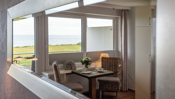 A dining area with a table set for two, large windows with a view of the sea, and a refrigerator in the background. - 1-room apartment with sea view and approx. 34 m² living space, for up to 2 people (Wenningstedt-Braderup (Sylt))