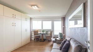 A modern living room with a sofa, a dining table with chairs, and a large window. - 1-room apartment with sea view and approx. 34 m² living space, for up to 2 people (Wenningstedt-Braderup (Sylt))