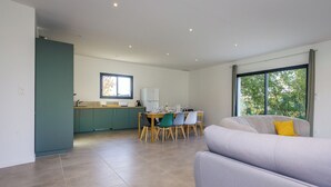 Een moderne keuken met blauwgroene kastjes, een eethoek met een tafel en stoelen, en een groot raam met uitzicht op bomen.