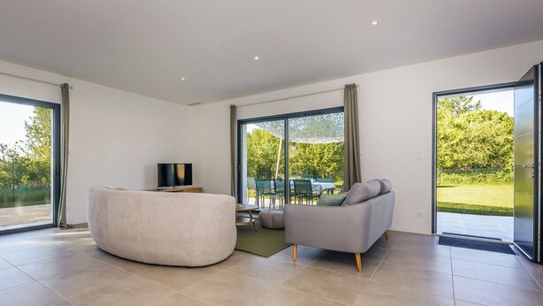 Een ruime woonkamer met een groot raam, een grijze bank, een rond houten poef en een flatscreen televisie op een tv-meubel.