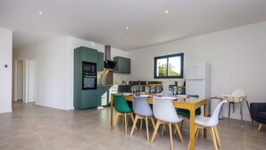 Een moderne keuken met blauwgroene kastjes, een eethoek met een houten tafel en stoelen, en uitzicht op de omgeving door een raam.