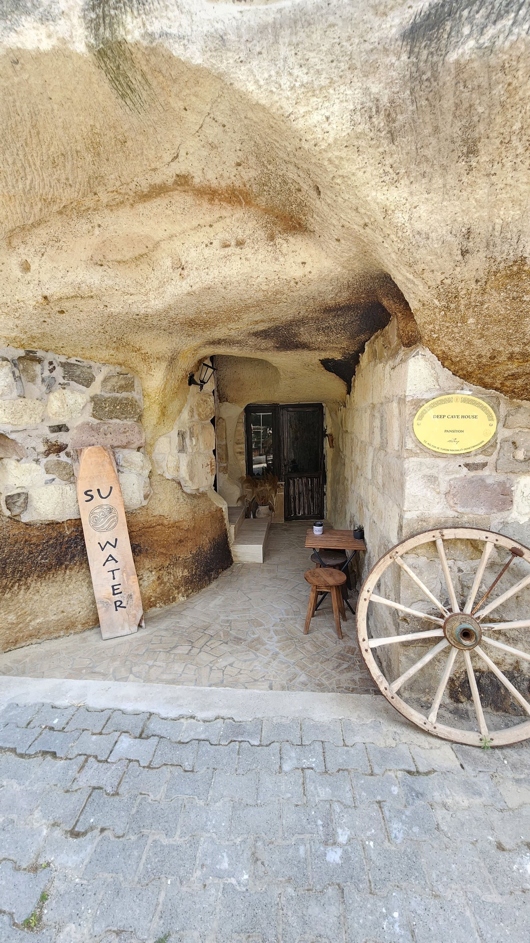 A stone entrance to a cave house with a wooden sign, a small table, and a large wooden wheel.