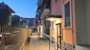 A narrow stone-paved pathway flanked by buildings with balconies, railings, and a metal fence.