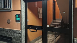 A hotel entrance with a glass door, a staircase inside, and a wall-mounted intercom.