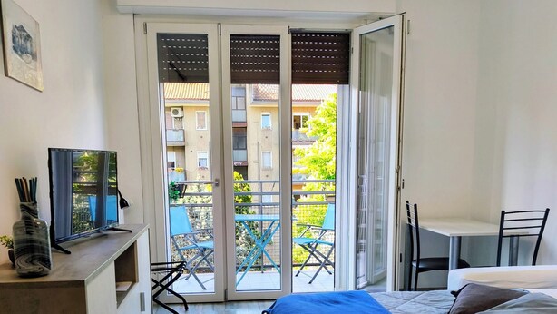A bedroom with a bed, a desk with a computer, a chair, and a balcony with a table and chairs.