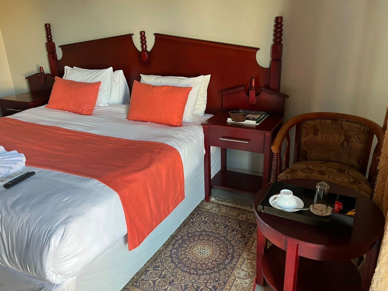 Habitación de hotel con una cama grande, cabecera de madera, mesita de noche, una silla y una pequeña mesa con bandeja que contiene una tetera y tazas.