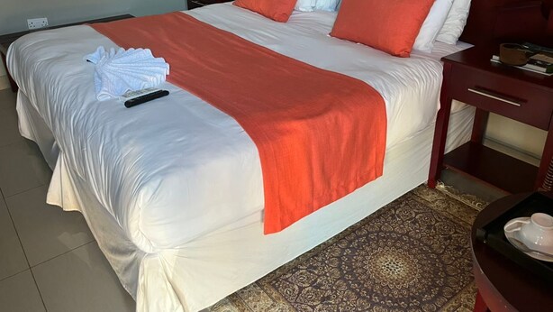A neatly made bed with white and orange bedding, a remote control, and a folded towel on the bed. - Kalimba Guesthouse (Gaborone)