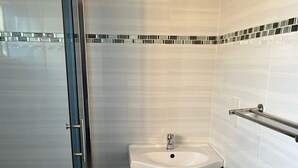 A bathroom with a glass shower enclosure, a white sink, dark cabinets, and a patterned floor. - Kalimba Guesthouse (Gaborone)