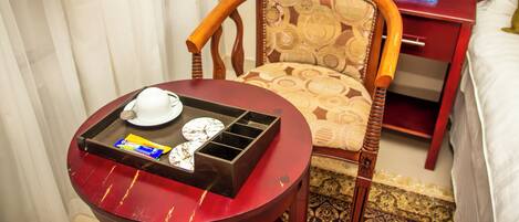 A hotel room with a wooden chair, a small table with a tray, a laptop on a red nightstand, and a bed with white bedding.