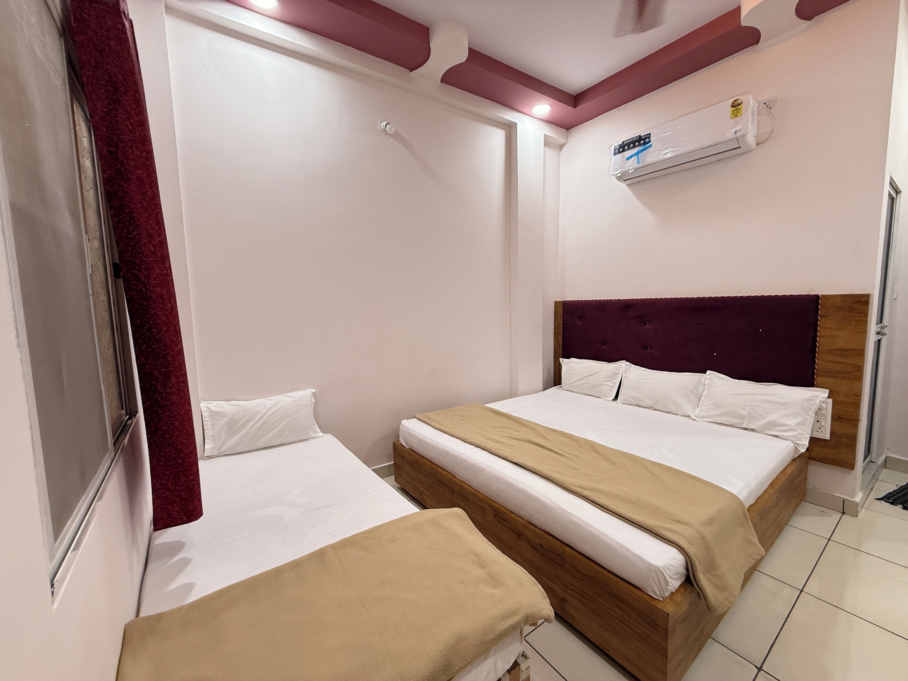 Habitación de hotel con dos camas, cabecero de madera, ventana con cortina roja y unidad de aire acondicionado en la pared.