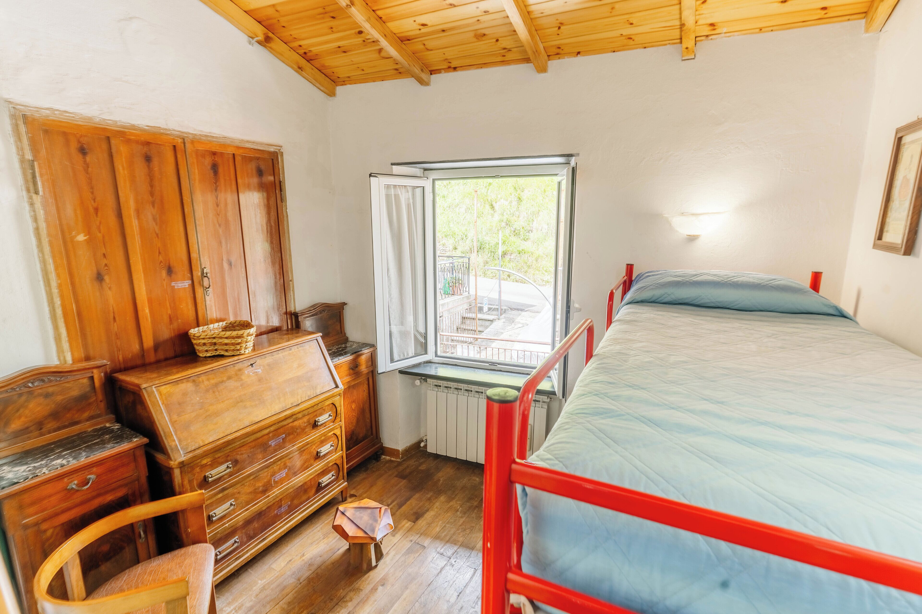 A bedroom with a wooden wardrobe, a bed with a red frame, a window with a view of greenery, and a small stool.