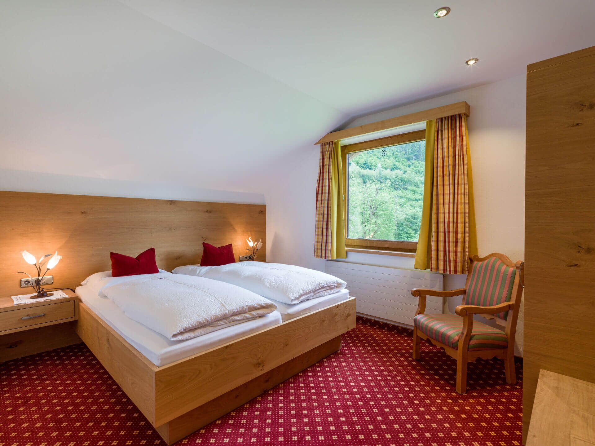 Un lit double avec deux oreillers rouges, une tête de lit en bois, une table de chevet avec une lampe, une fenêtre avec des rideaux, une chaise en bois et un tapis rouge à motifs.