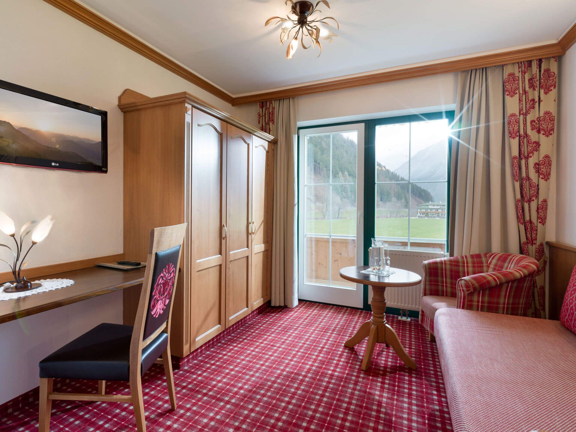 A hotel room with a checkered red carpet, a wooden wardrobe, a desk with a chair, a bed, a table with a chair, and a large window with a view of mountains.