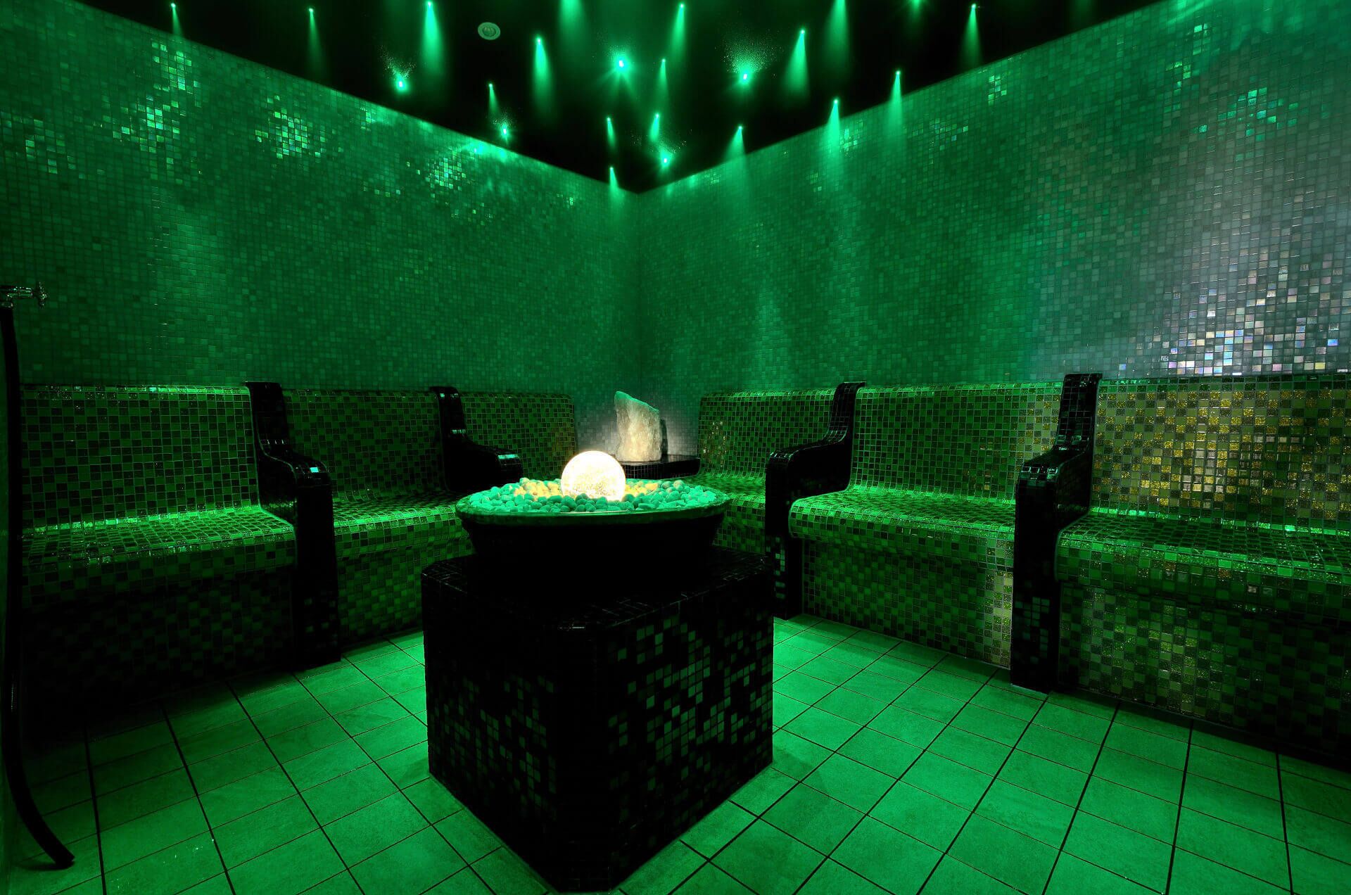 A spa room with a central heated stone feature, green ambient lighting, and tiled walls and floor.