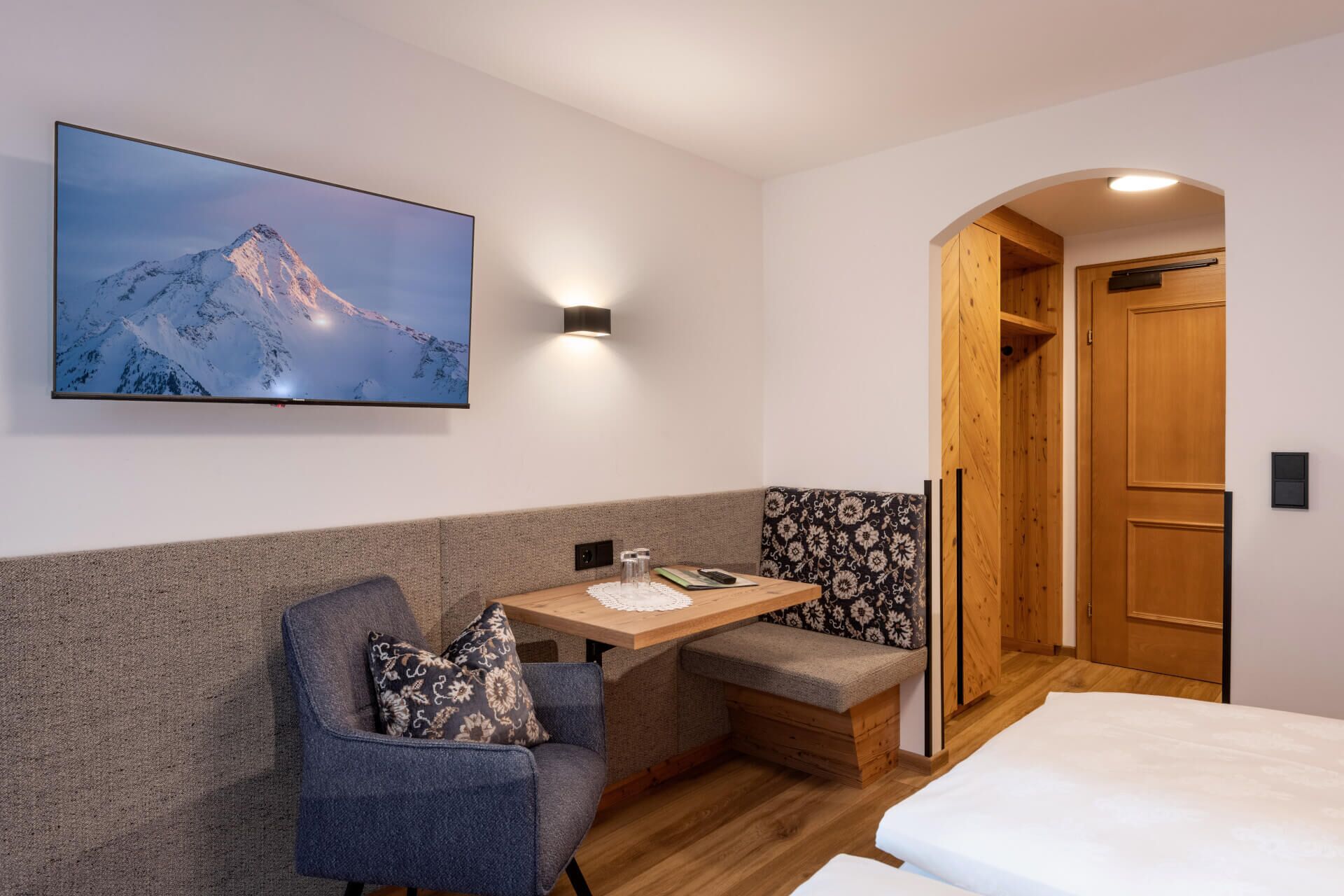 A cozy room with a bed, a seating area with a table, a wall-mounted TV, and a wooden wardrobe.