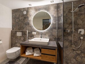 A modern bathroom with a stone-patterned wall, a round mirror, a sink, a shower, and a toilet. - Ferienhof Oblasser (Mayrhofen)