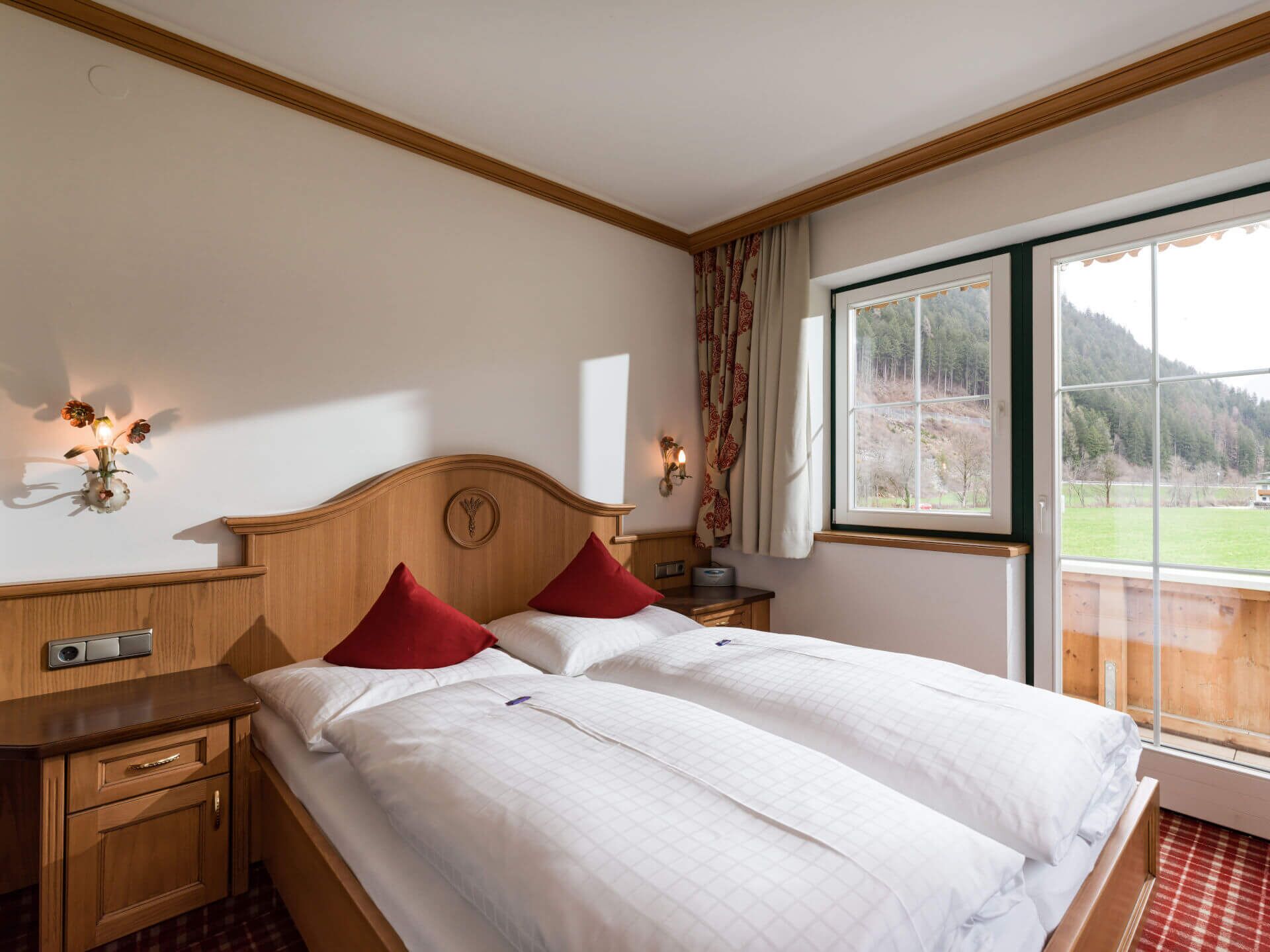 A double bed with red pillows, a wooden headboard, a nightstand with a lamp, and a window with a view of a forest.