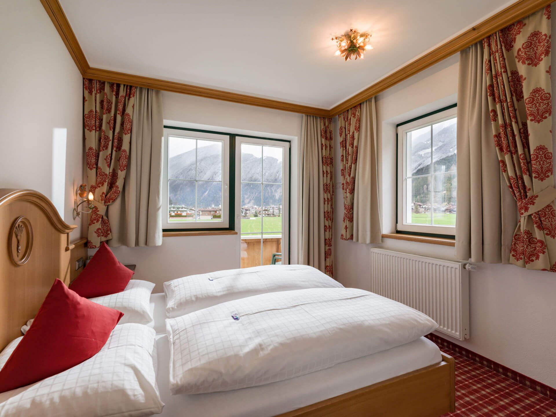 A hotel room with a bed, two red pillows, a wooden headboard, two windows with curtains, and a view of a mountain and town.