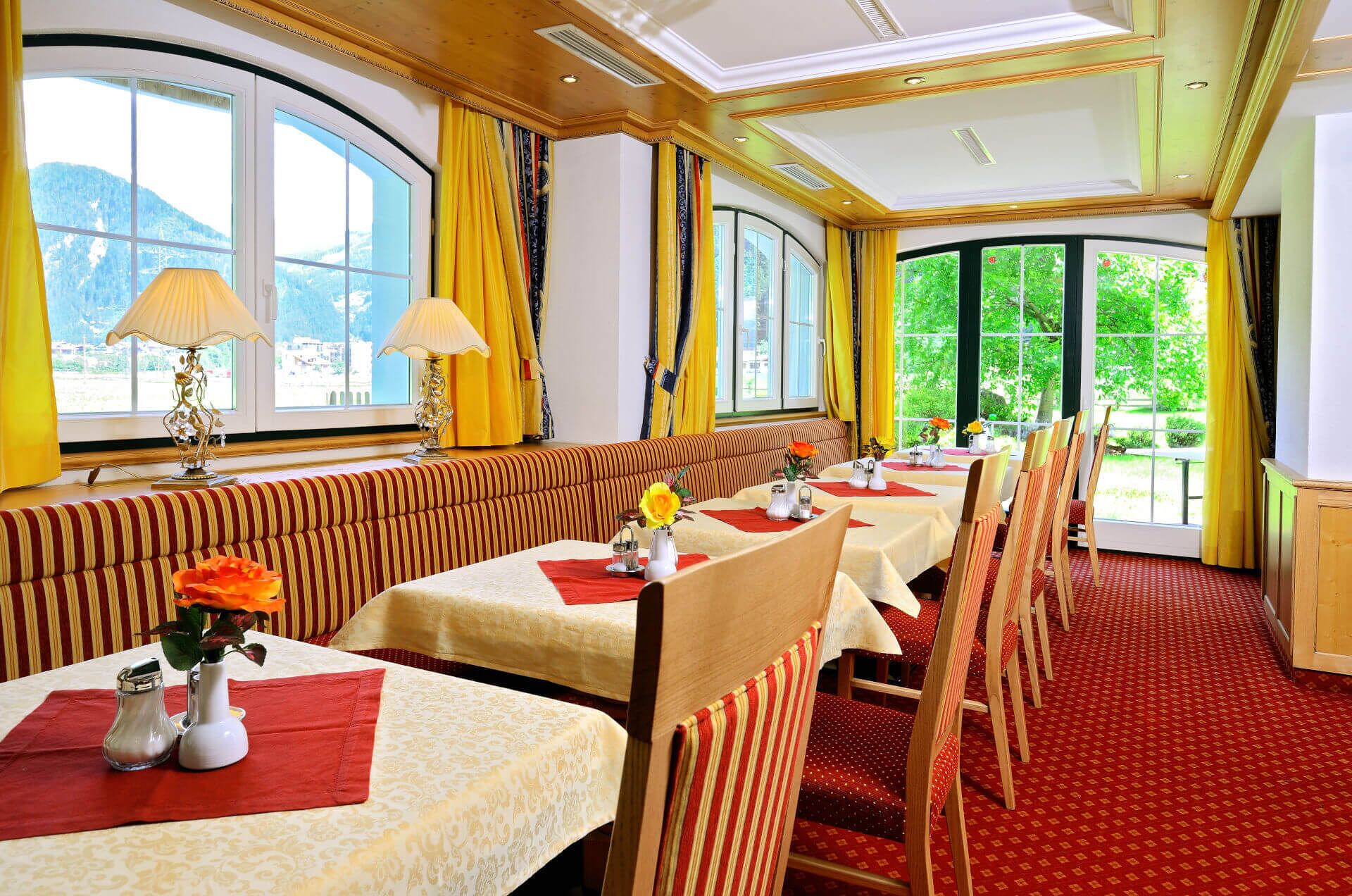 A dining area with tables set for four, a striped bench, and large windows with curtains.