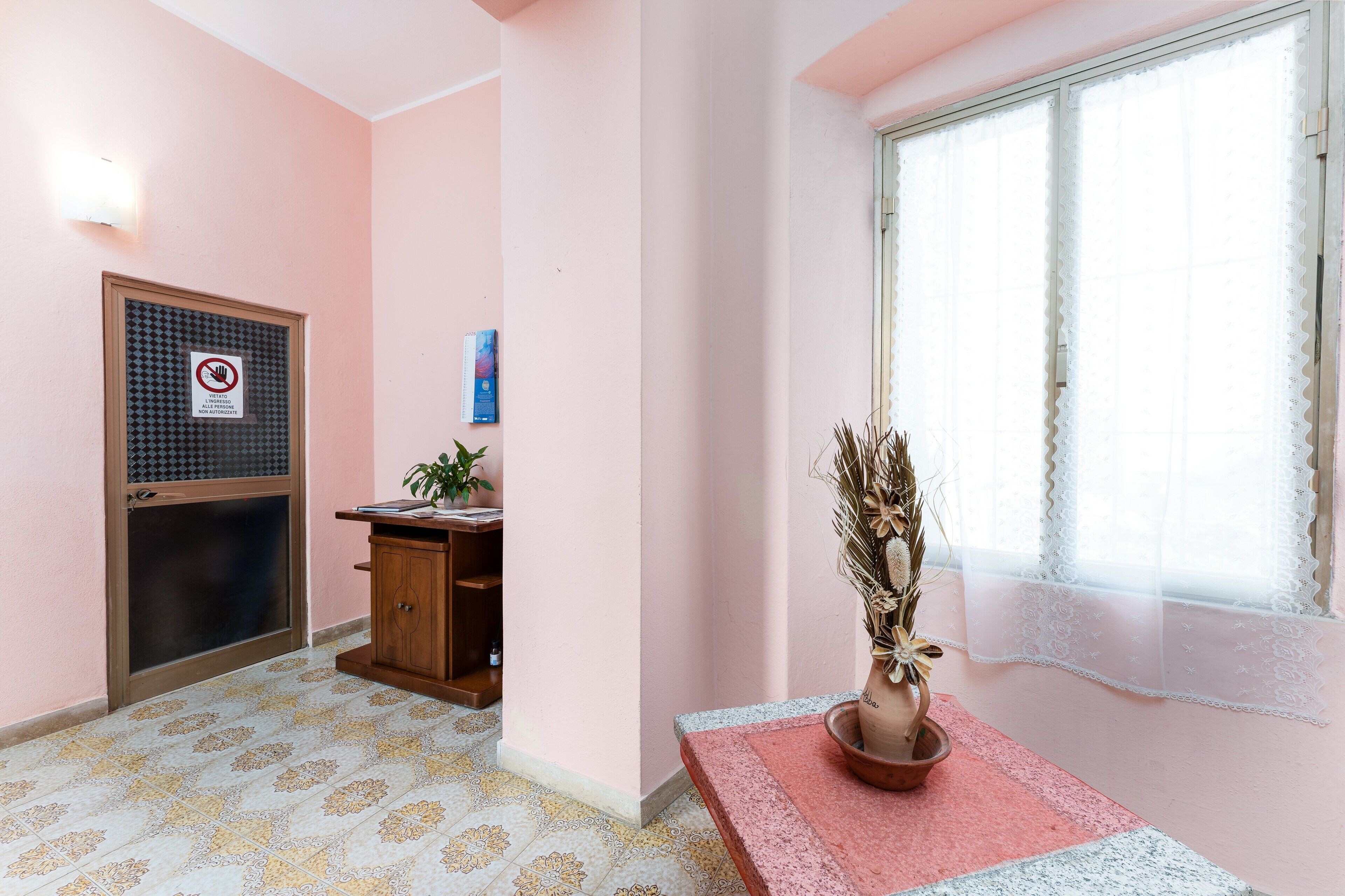 Una habitación con pared rosa, una puerta con mosquitero, un pequeño armario de madera, una planta en maceta y una ventana con cortinas de gasa.