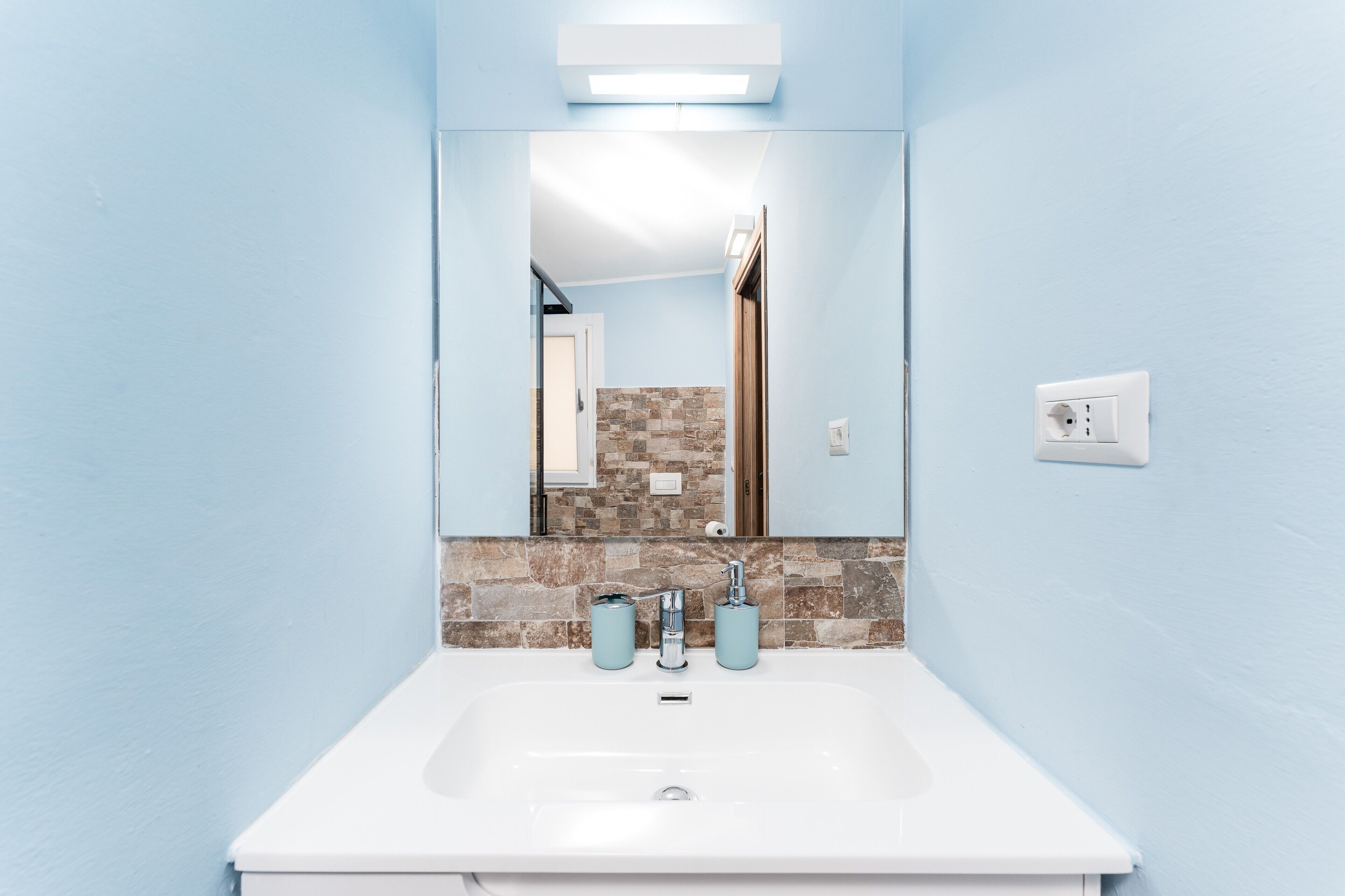 Een badkamer met een witte wastafel, twee lichtblauwe zeepdispensers, een spiegel en een wandlamp.