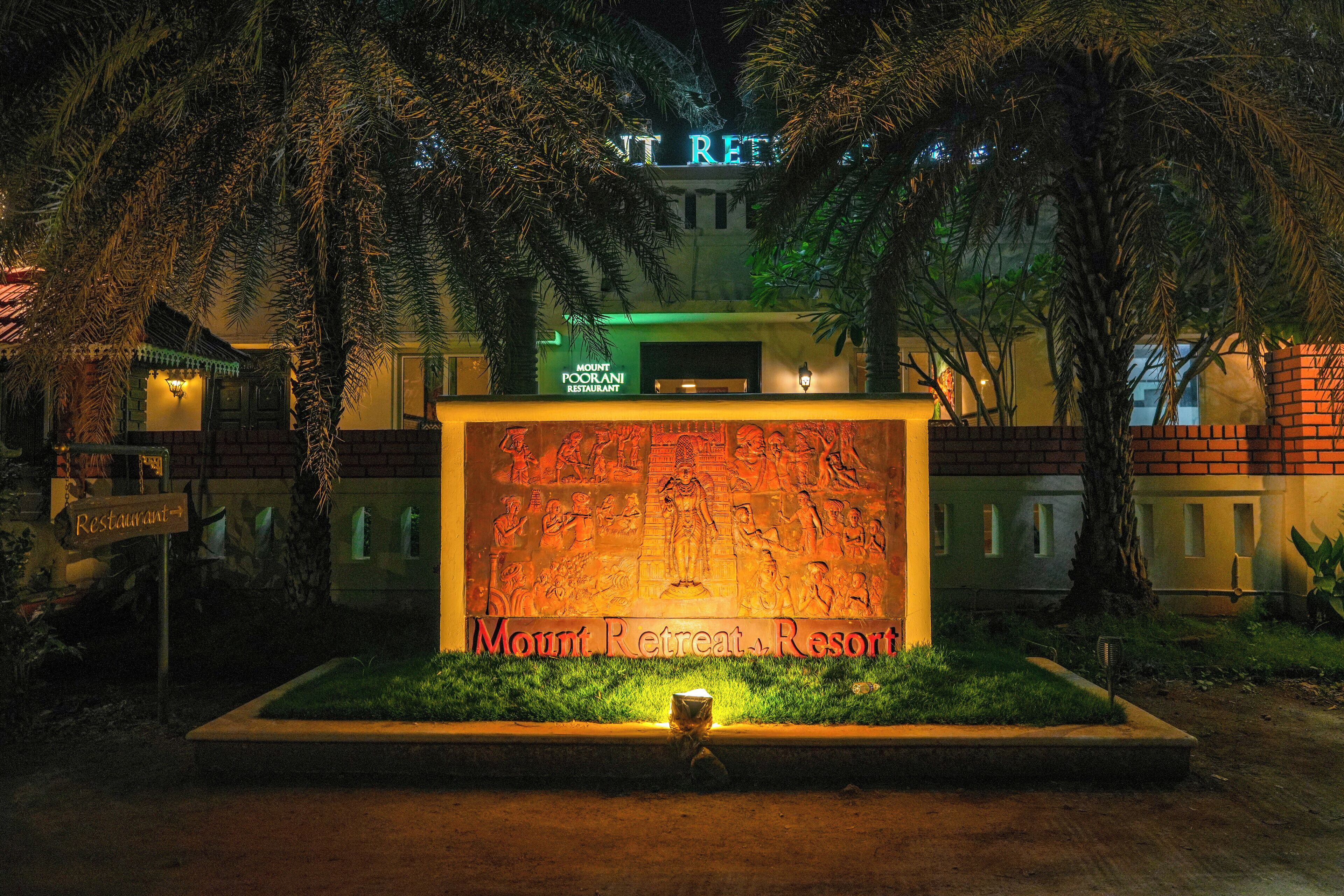 Entree van een resort met een groot verlicht bord, palmbomen en een bakstenen muur.