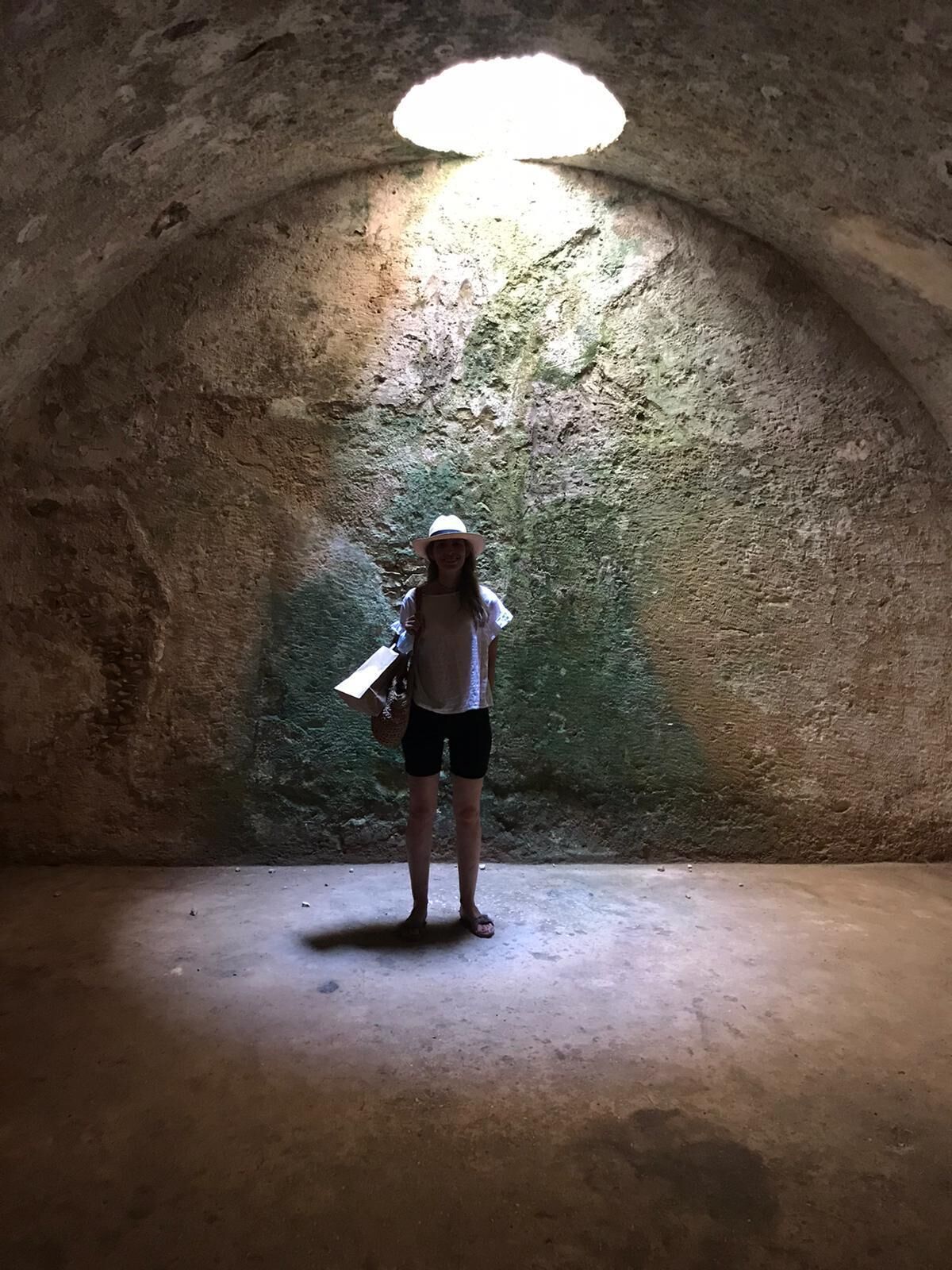 A person standing in a dimly lit, arched stone room with a single light source at the top.