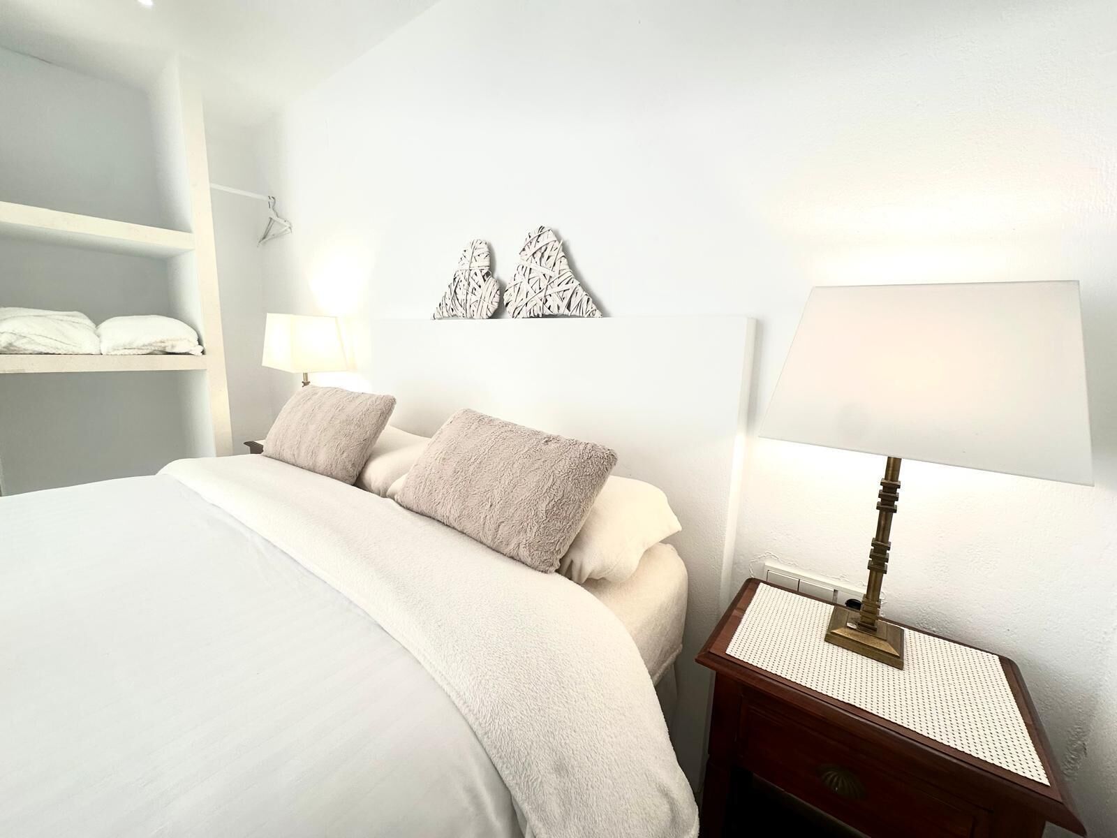 Une chambre à coucher bien rangée, avec un lit, deux oreillers, une table de chevet avec une lampe et une étagère fixée au mur où sont pliées des serviettes.