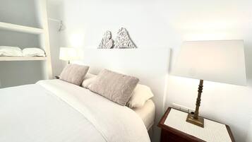 Une chambre à coucher bien rangée, avec un lit, deux oreillers, une table de chevet avec une lampe et une étagère fixée au mur où sont pliées des serviettes.