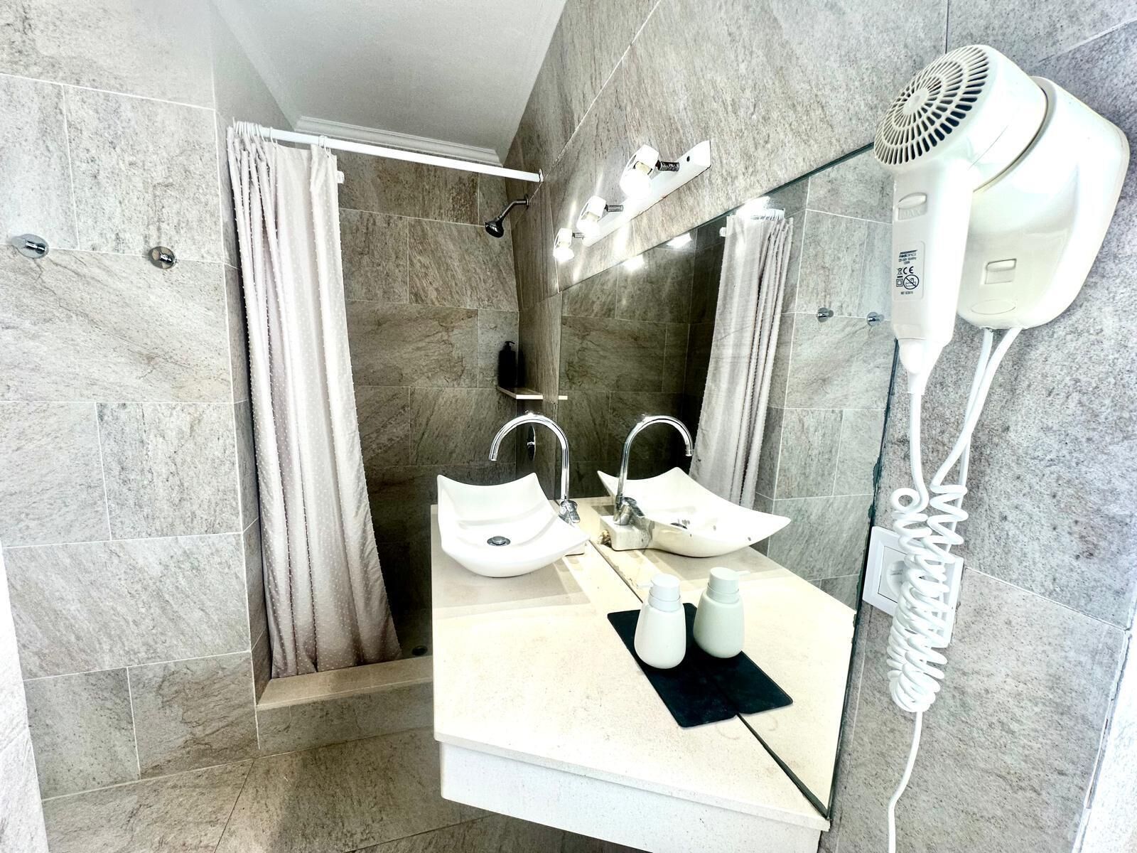A modern bathroom with a large mirror, two sinks, a shower area, and a hairdryer mounted on the wall.