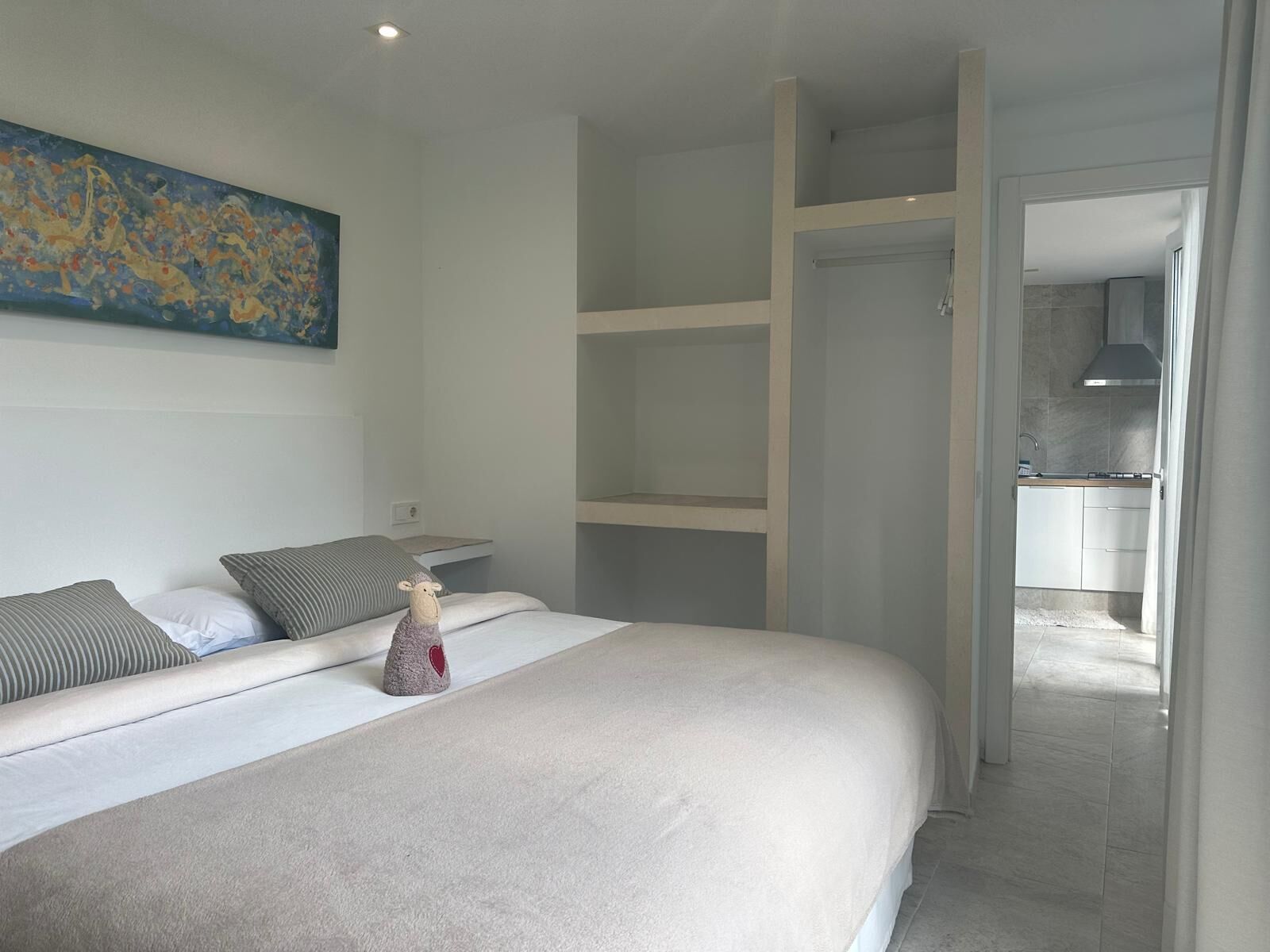 A modern bedroom with a large bed, a painting on the wall, and a view into the kitchen.