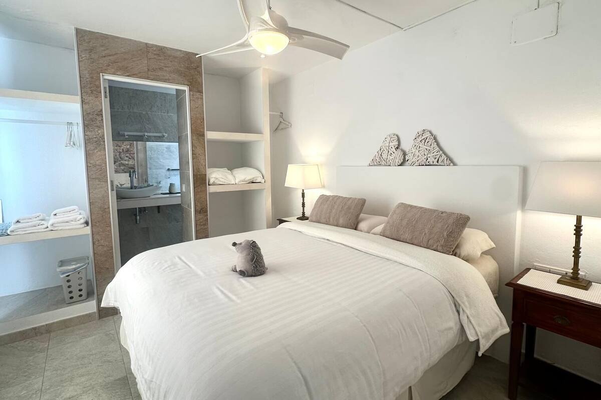Un dormitorio con cama, mesita de noche, lámpara, estante en la pared, perchero y ventilador de techo.