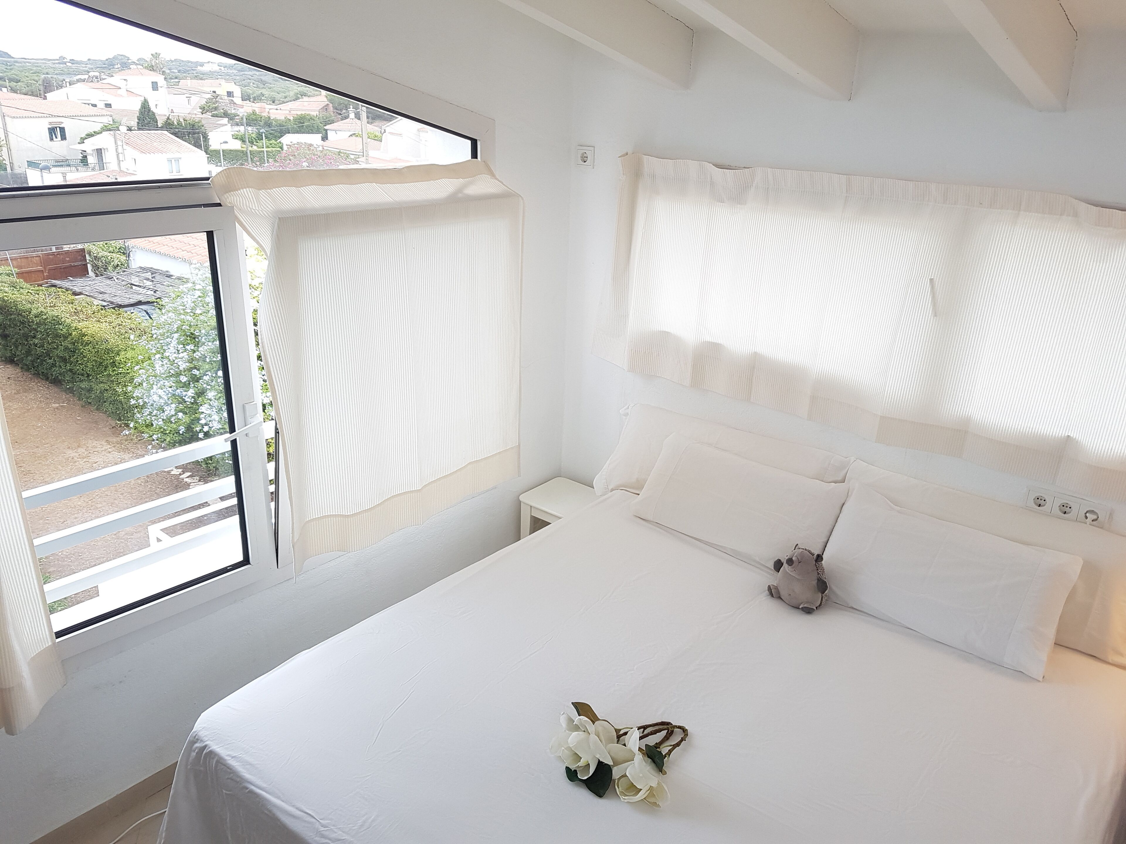 A bedroom with a large window, white bedding, and a small bedside table.