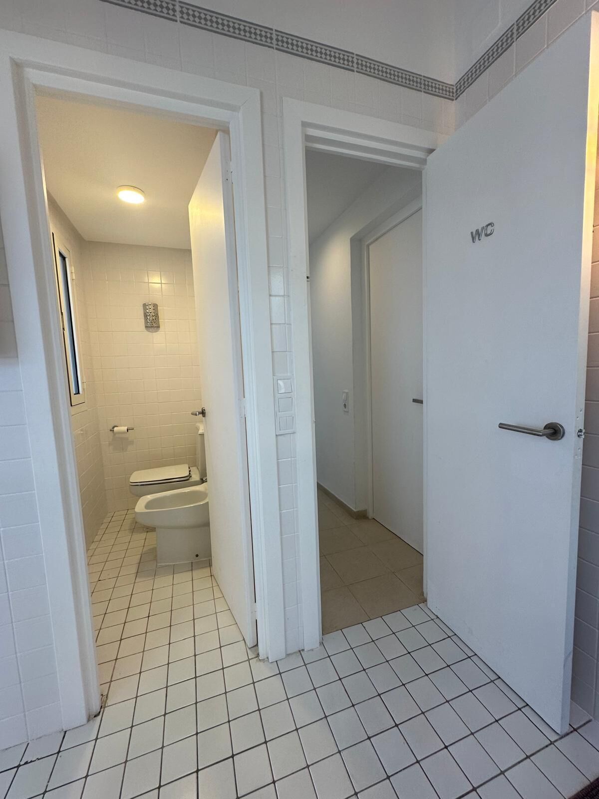 A bathroom with white tiled walls and floor, a toilet, and a shower.