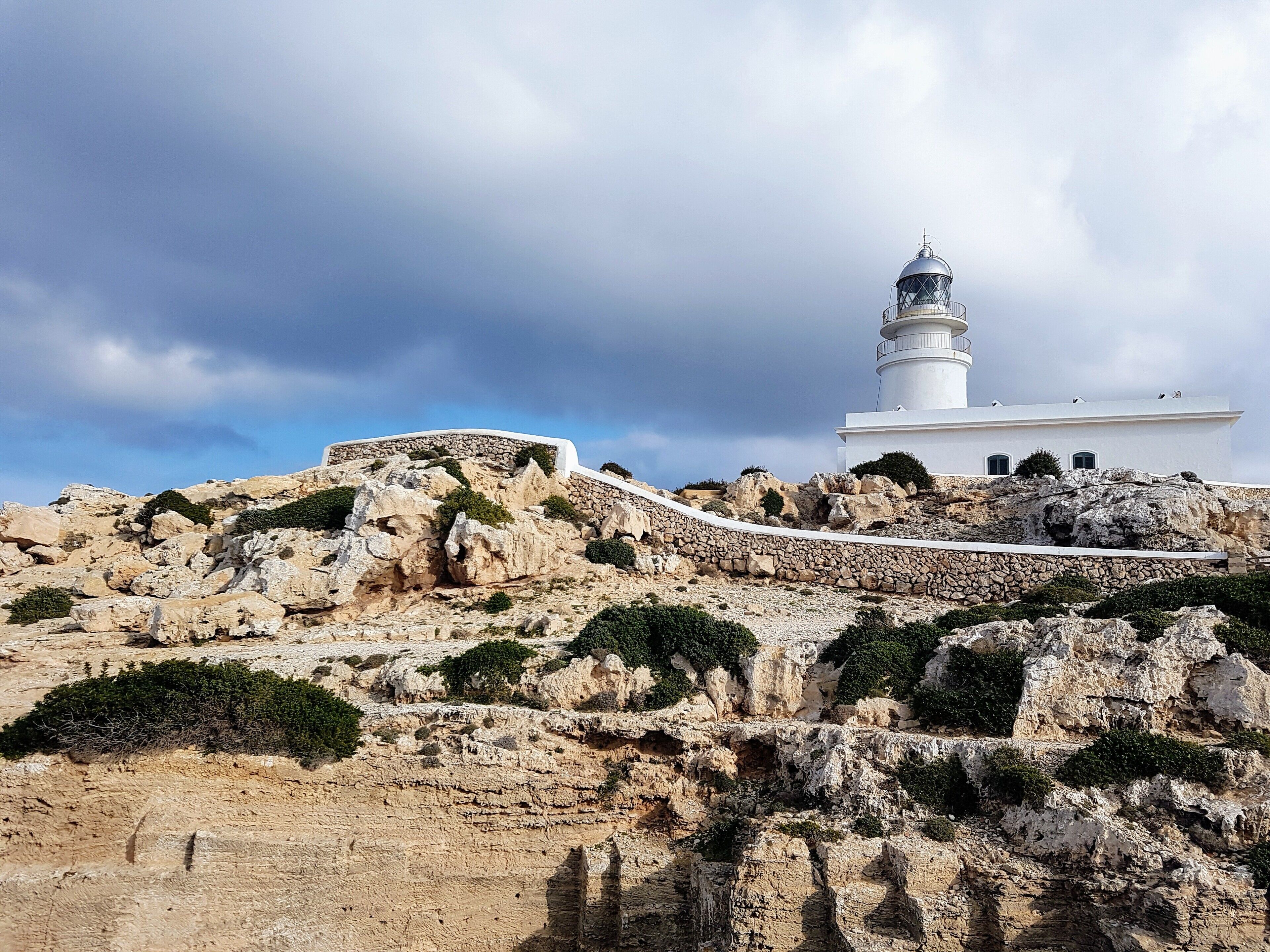 A white lighthouse on a rocky hill with a stone wall and vegetation.