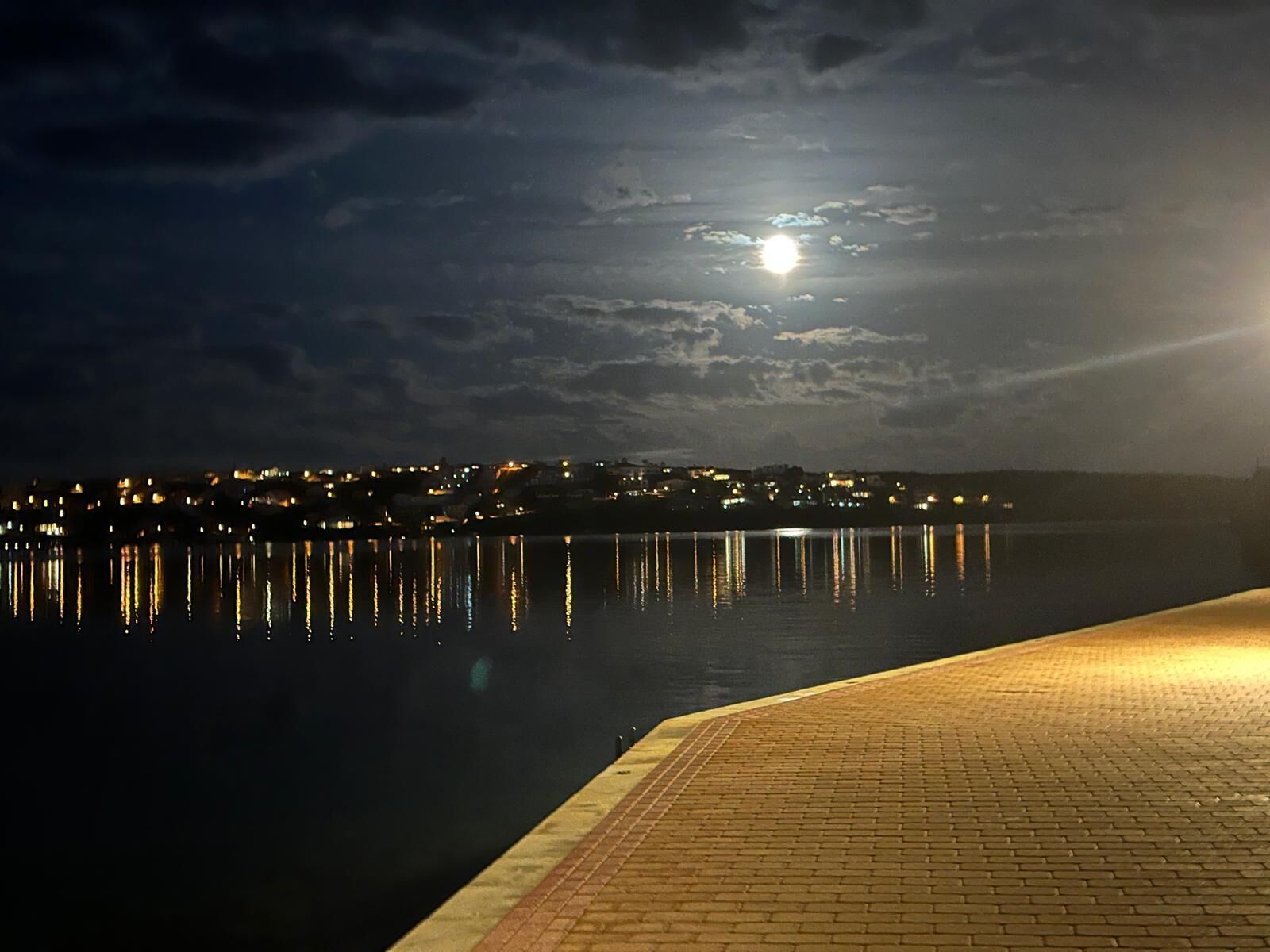 A nighttime waterfront view with a paved walkway and city lights reflecting on the water.