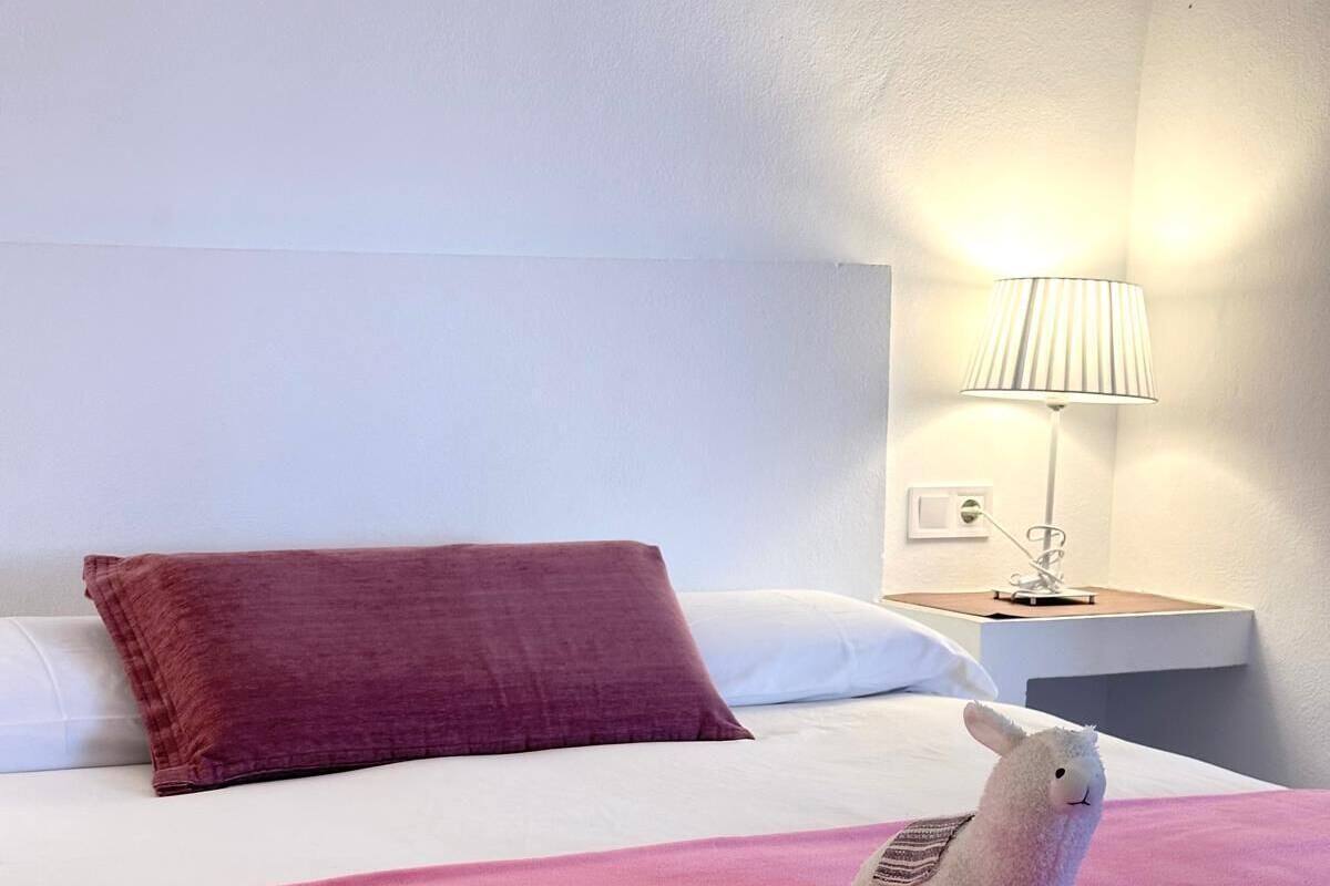 Una cama bien hecha con un edredón rosa, una almohada roja y una almohada blanca. Hay una lámpara pequeña sobre una mesita de noche junto a la cama.