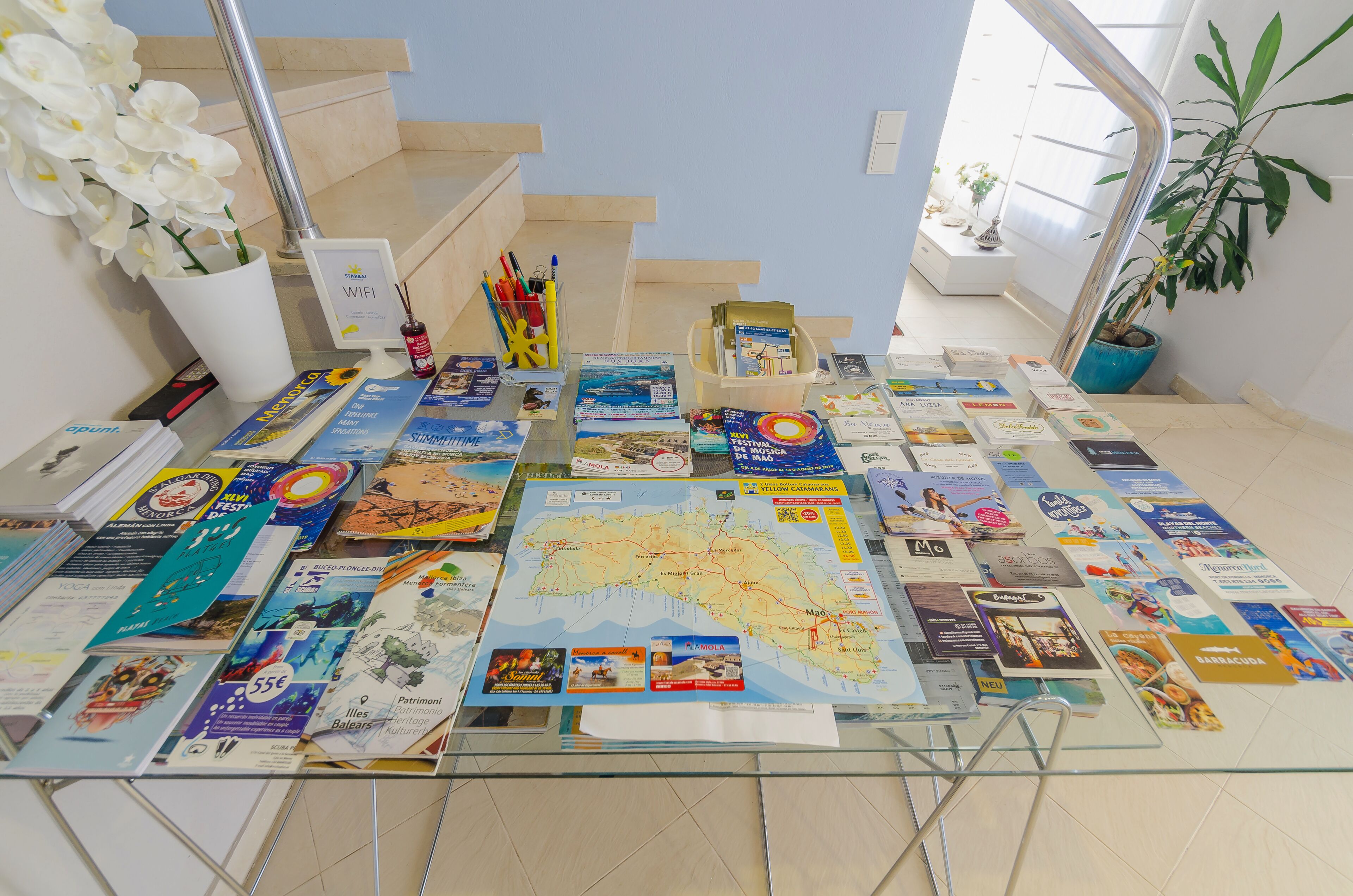 A glass table with various travel brochures and a map, surrounded by a staircase and potted plants.