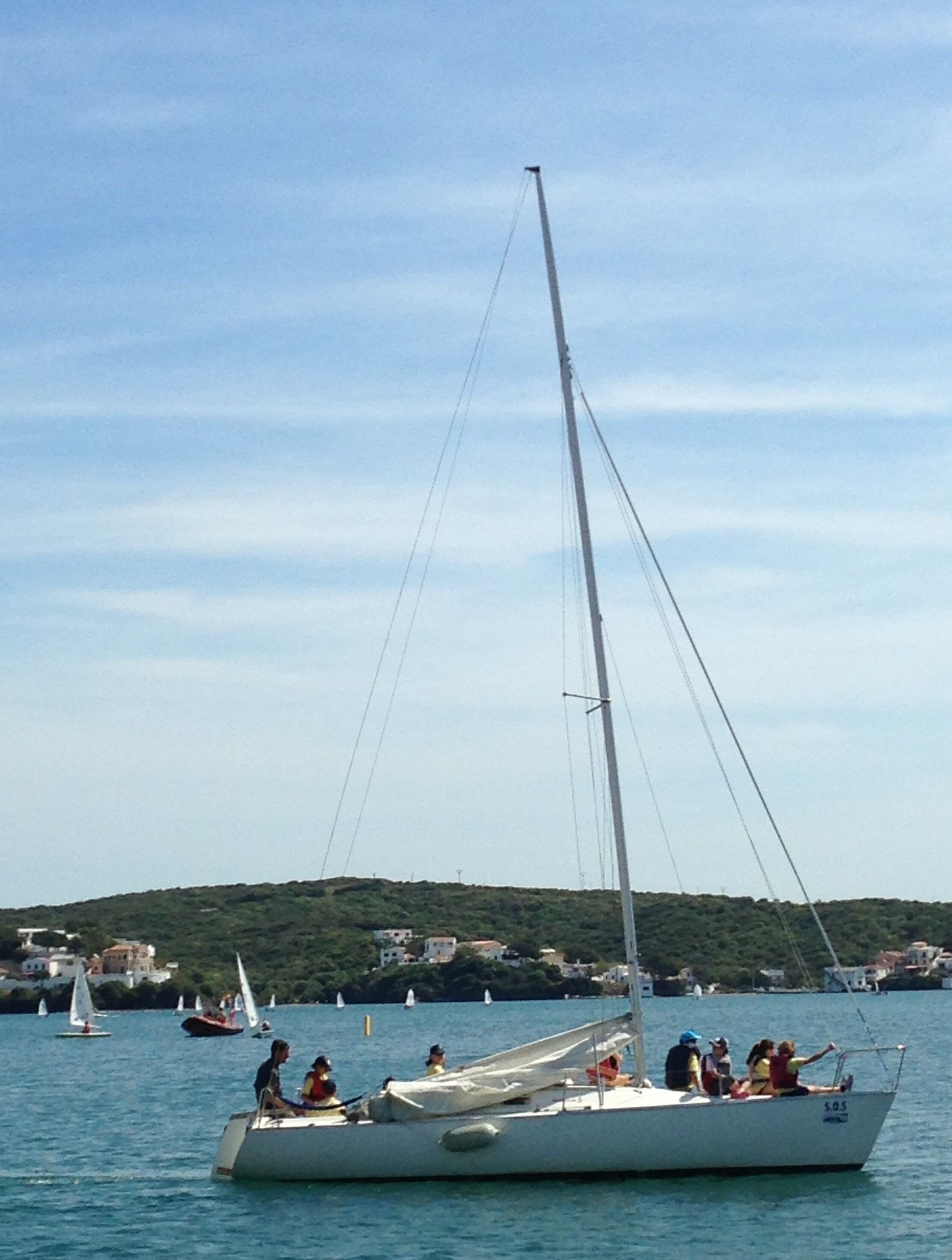 Un velero con pasajeros, amarrado cerca de una zona costera con edificios al fondo.