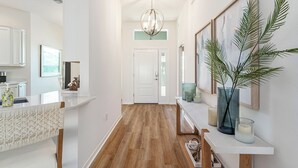 Un couloir doté d’un plancher en bois, de murs blancs, d’une porte blanche, d’un coin cuisine avec des meubles blancs et d’une étagère ornée d’objets décoratifs.