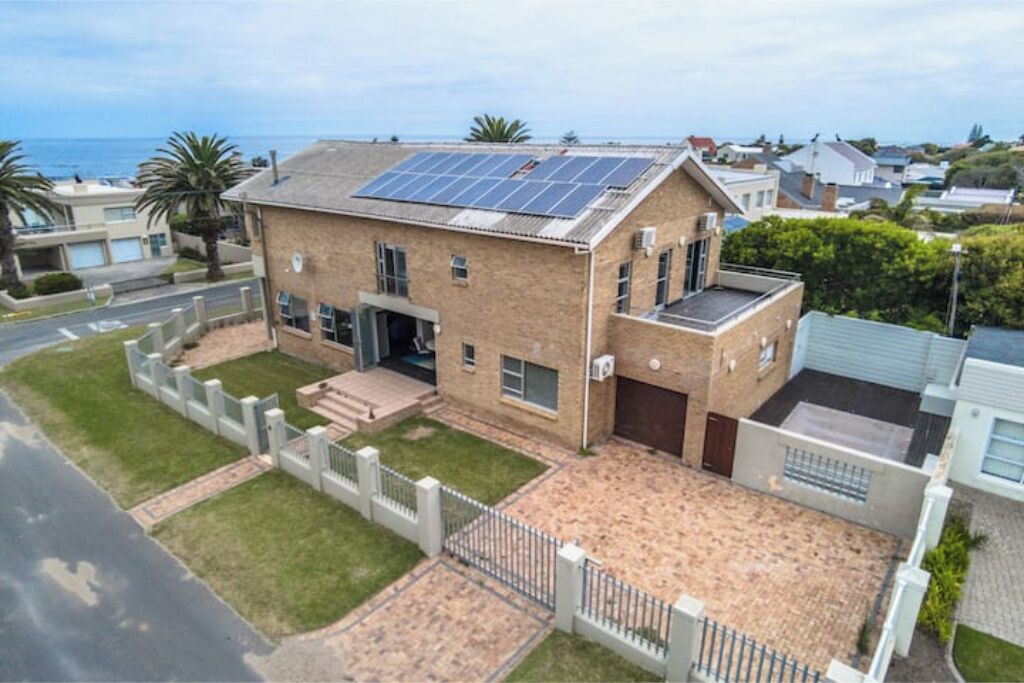 A two-story house with a solar panel roof, a fenced yard, and a view of the ocean.