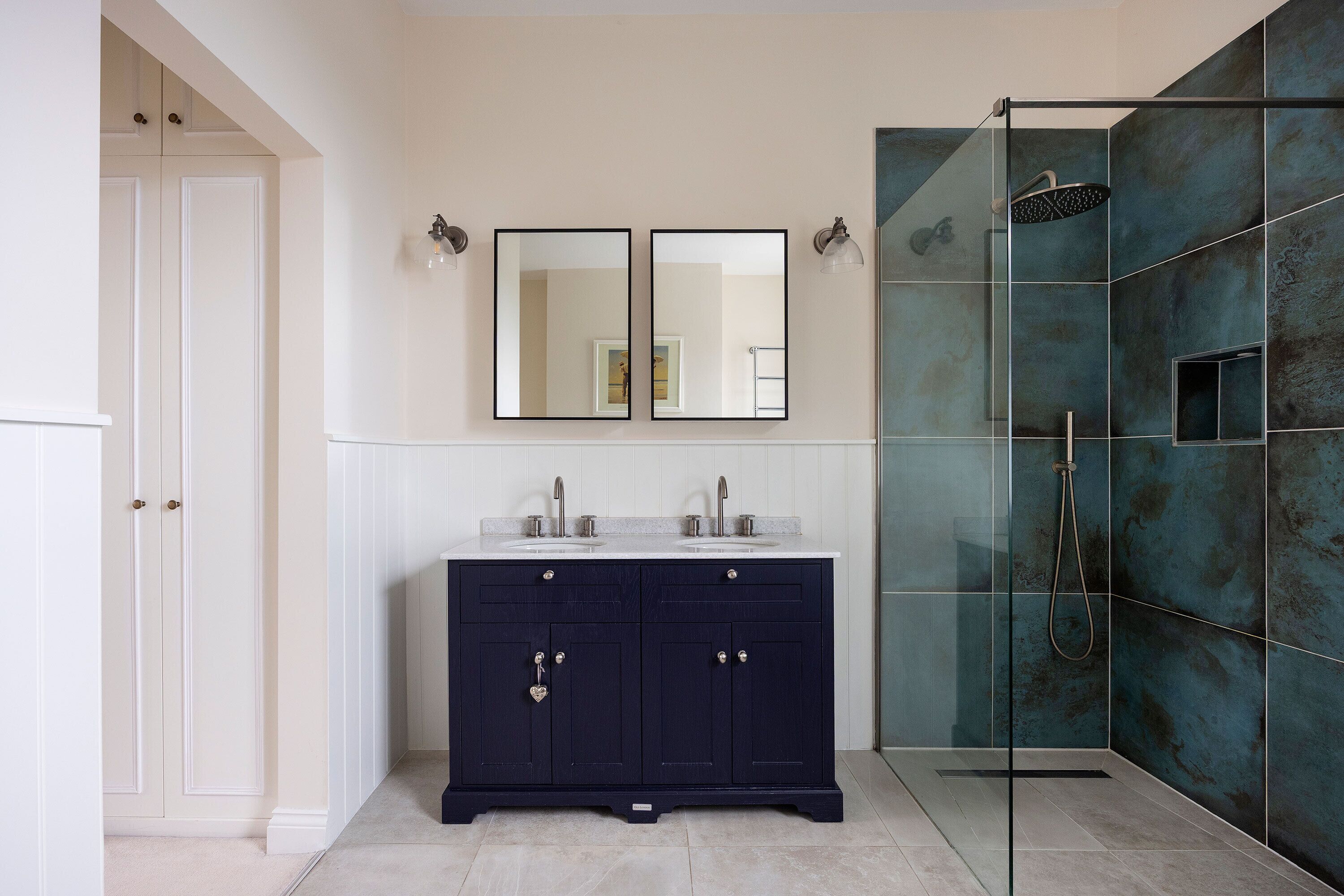Une salle de bain avec un meuble-lavabo bleu foncé, deux miroirs, une cabine de douche en verre et une porte blanche.