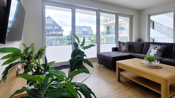 Een moderne woonkamer met grote ramen, een bank, een salontafel en potplanten.