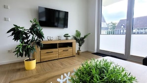 Een moderne woonkamer met een houten vloer, een flatscreen televisie aan de muur en verschillende plantenpotten.
