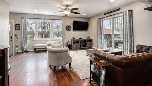 A spacious living room with hardwood flooring, a ceiling fan, a large window with curtains, a fireplace, and a leather sofa.