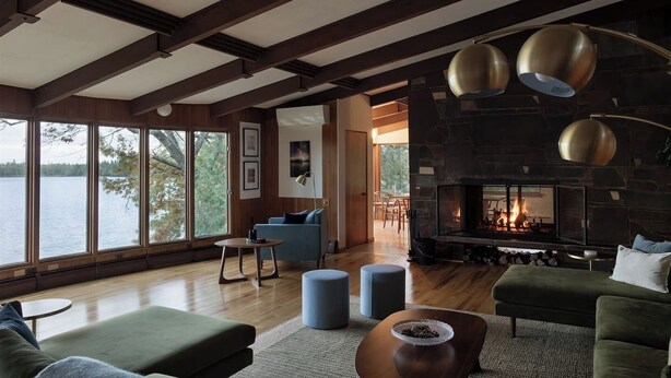Amplia sala de estar con chimenea de piedra, ventanas amplias con vista a un cuerpo de agua y muebles modernos.