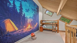 Un dormitorio con una litera, un tapiz de gran tamaño con motivos forestales, una pequeña decoración de hoguera y una ventana que da a vistas de árboles.