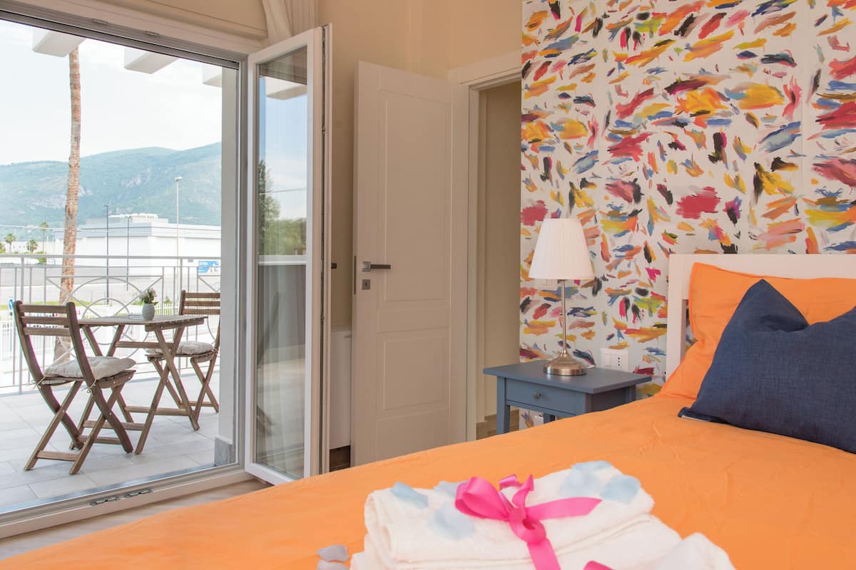 A hotel room with a bed, a window with a view of the outdoors, a door, and a small table with a lamp.